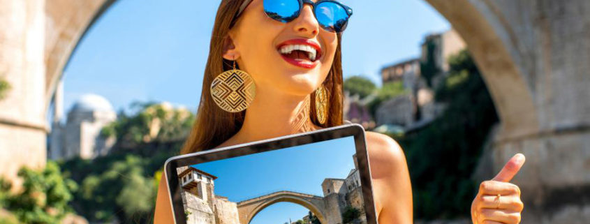 Une jeune femme travaillant pour un office de tourisme, souriante, tient dans sa main une tablette où apparaît un pont. Ce même pont est justement derrière elle, sous le soleil.