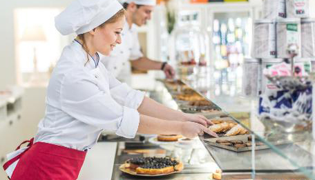 Une serveuse et un serveur, tous deux souriant, sont à l’action dans une pâtisserie et mettent en vitrine tartes et gâteaux.