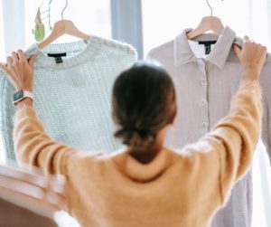 Dans un magasin de prêt-à-porter, une cliente, de dos, tend deux chemises devant elle pour les comparer à la lumière émanant d’une fenêtre.