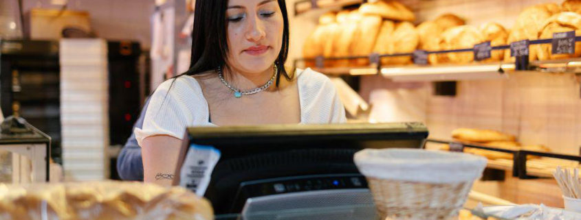 Une commerçante, dans sa boulangerie, semble se demander, devant sa caisse, quel logiciel de caisse PrestaShop choisir pour son commerce.