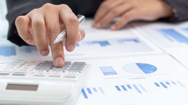 Main d’un commerçant posant le doigt sur une calculatrice. Des statistiques et rapports comptables sont étalés sur son bureau.