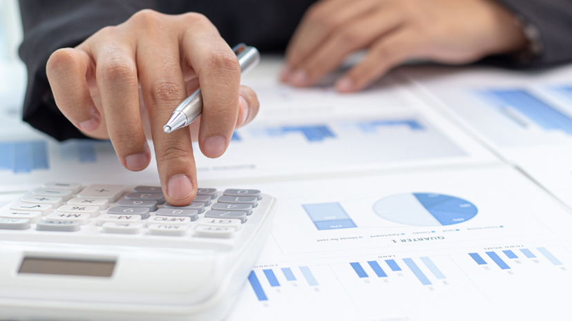 Main d’un commerçant posant le doigt sur une calculatrice. Des statistiques et rapports comptables sont étalés sur son bureau.