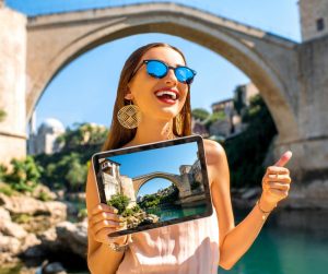 Une jeune femme en tenue estivale, près d’un très ancien pont, tient dans sa main une tablette utilisant le logiciel de caisse pour office de tourisme KerAwen.