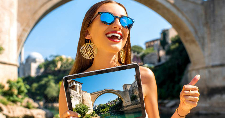 Une jeune femme travaillant pour un office de tourisme, souriante, tient dans sa main une tablette où apparaît un pont. Ce même pont est justement derrière elle, sous le soleil.