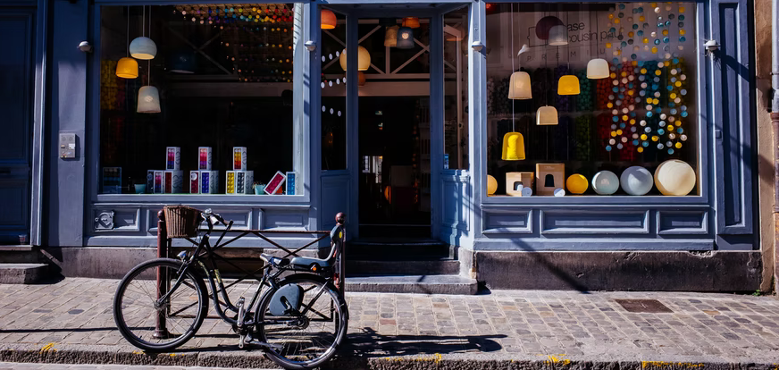 Vieux vélo appuyé contre un trottoir devant la devanture d'un concept store. À travers la vitrine, sont visibles des luminaires très tendance.