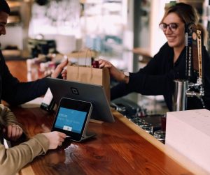 Transformez votre tablette en caisse enregistreuse, comme cette commerçante au comptoir de son magasin, qui remet à un couple ses achats.